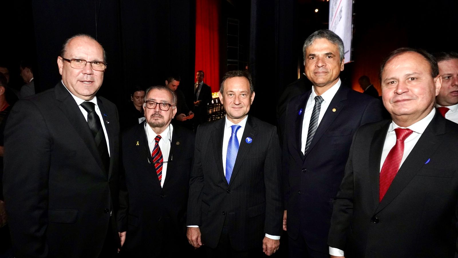 Esta é uma fotografia que mostra cinco homens, todos vestidos formalmente, lado a lado em um evento social ou de negócios. Todos usam terno escuro. Eles posam juntos em um ambiente interno escuro.