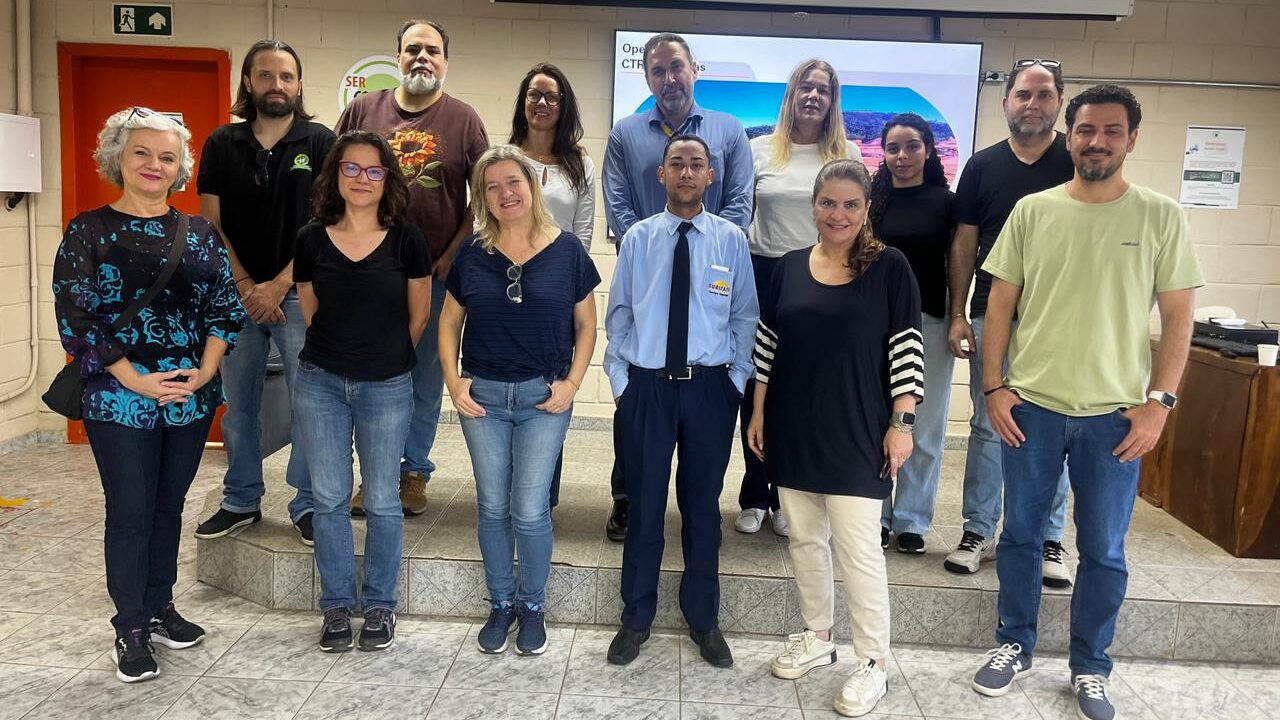 Foto de grupo em um ambiente interno, posando em dois níveis (primeiro plano no chão e segundo plano em um pequeno tablado).

O grupo de pessoas estão vestidas com roupas casuais (jeans e camisetas/blusas), com exceção de um homem no centro, que veste uniforme social (calça e camisa social azul clara com crachá).