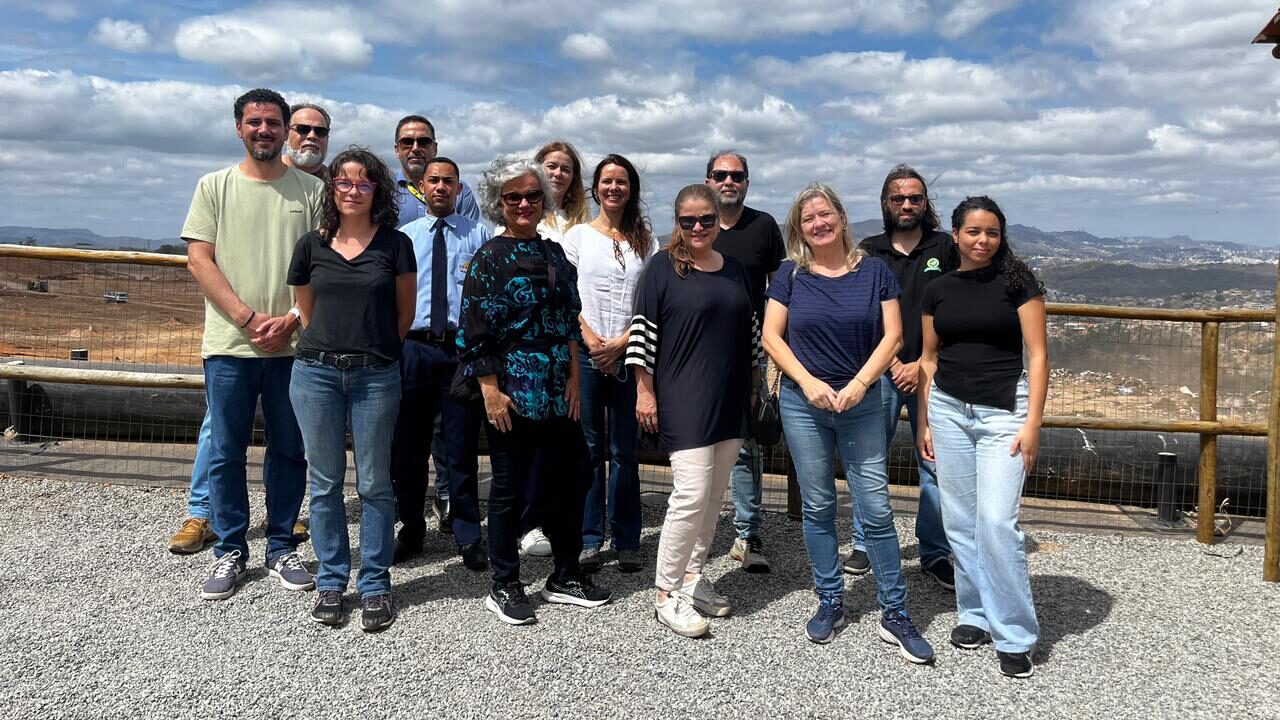Foto de um grupo de pessoas em pé, posando para a câmera.

O grupo está vestido com roupas casuais (camisetas, camisas polo, jeans e tênis), e a maioria está usando óculos de sol. Eles estão sobre um piso de pedras.

O fundo da imagem é uma vista panorâmica de uma paisagem urbana/rural distante, sob um céu parcialmente nublado.