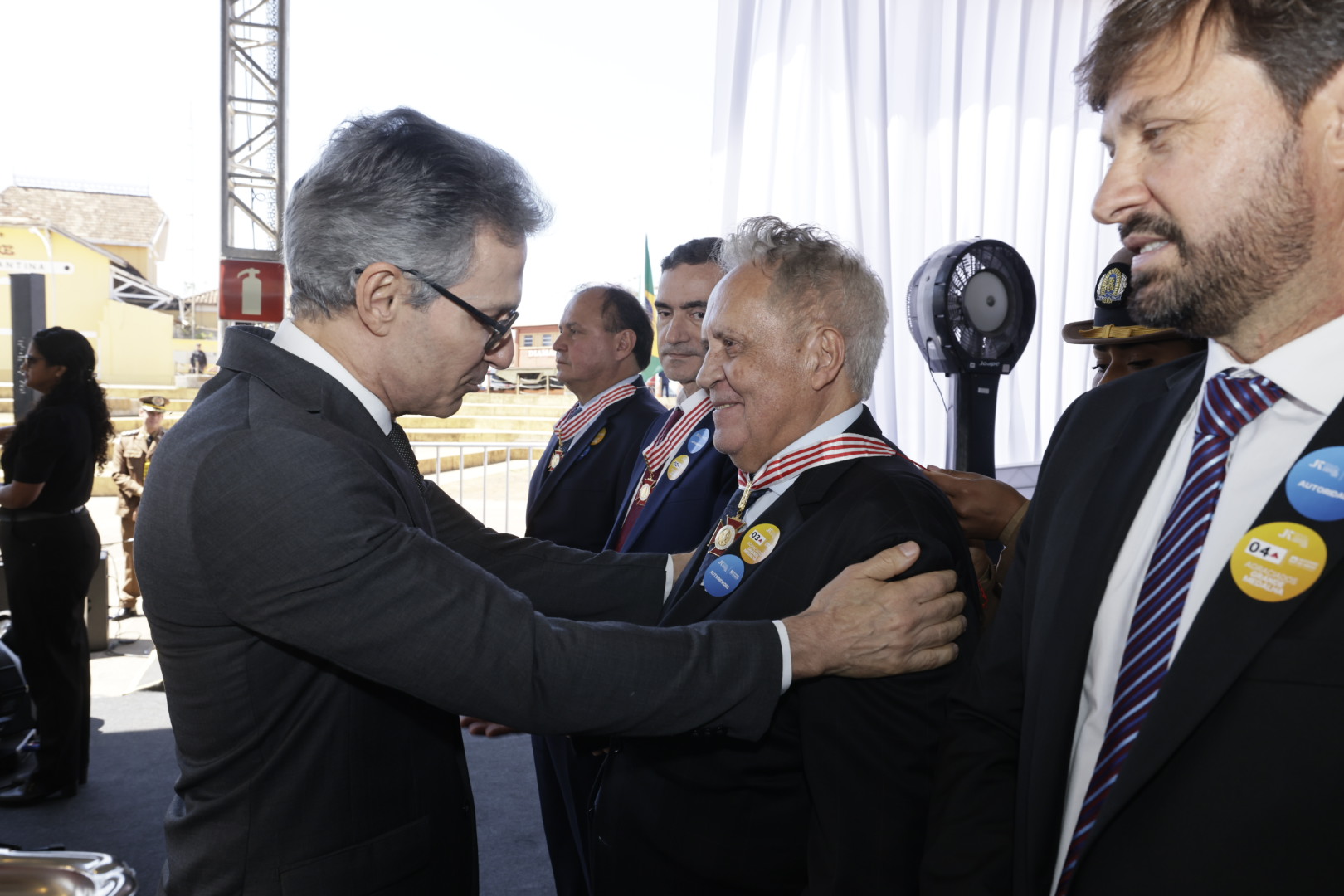 Um homem de óculos e terno escuro parabeniza outro, em uma cerimônia solene.