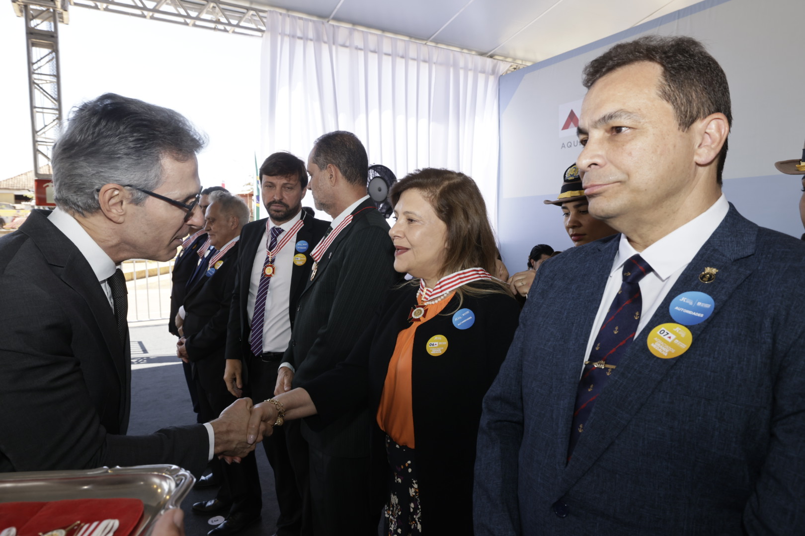 Um homem de terno e óculos aperta a mão de uma mulher durante uma cerimônia. Várias pessoas, incluindo homens de terno e uma mulher com medalhas no pescoço, estão na fila.