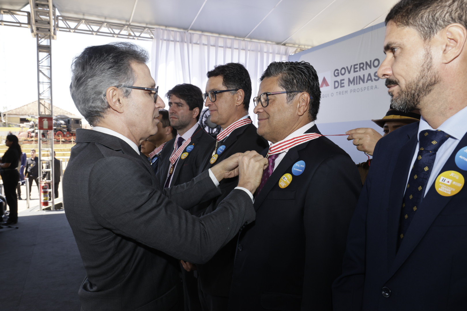 Um homem de óculos e terno escuro ajusta a medalha no pescoço de outro homem, que sorri, durante uma cerimônia solene ao ar livre, com um banner do "Governo de Minas" no fundo.