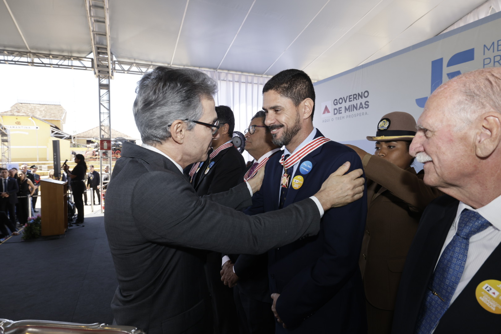 Um homem de óculos e terno escuro parabeniza outro, que veste terno e uma medalha, em uma cerimônia externa, com um banner do "Governo de Minas" ao fundo.