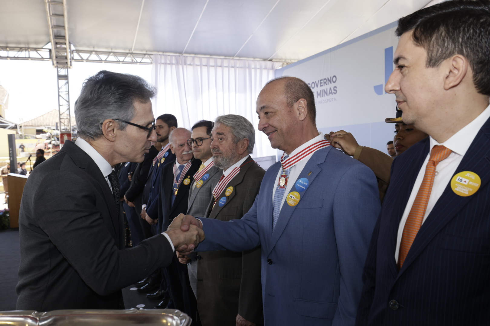 Dois homens de terno se cumprimentam com um aperto de mãos durante uma cerimônia solene ao ar livre. O homem da direita usa uma medalha no pescoço.