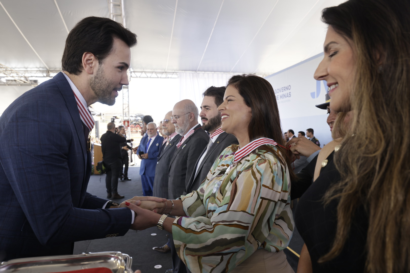 Um homem de terno azul-marinho cumprimenta uma mulher de blusa estampada e saia bege em uma cerimônia solene. Outras pessoas de terno estão enfileiradas ao fundo.