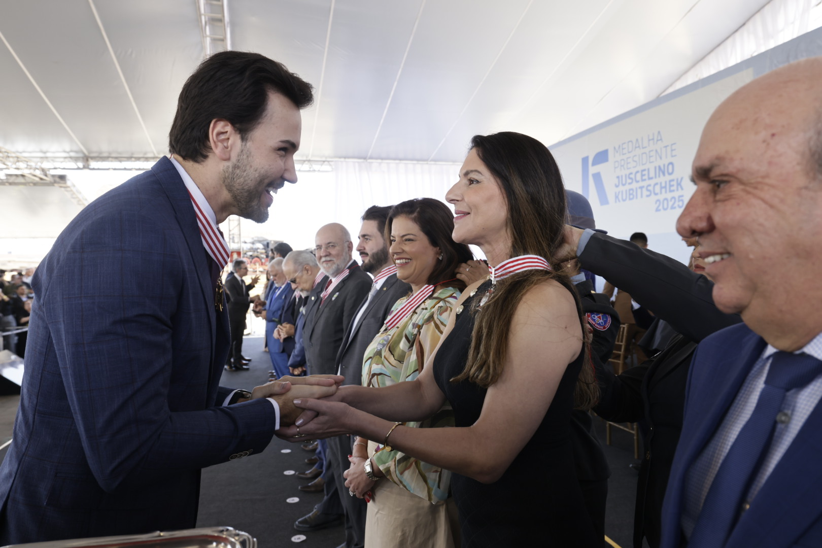 Um homem de terno azul-marinho sorri enquanto cumprimenta com um aperto de mãos uma mulher de vestido preto e medalha no pescoço.