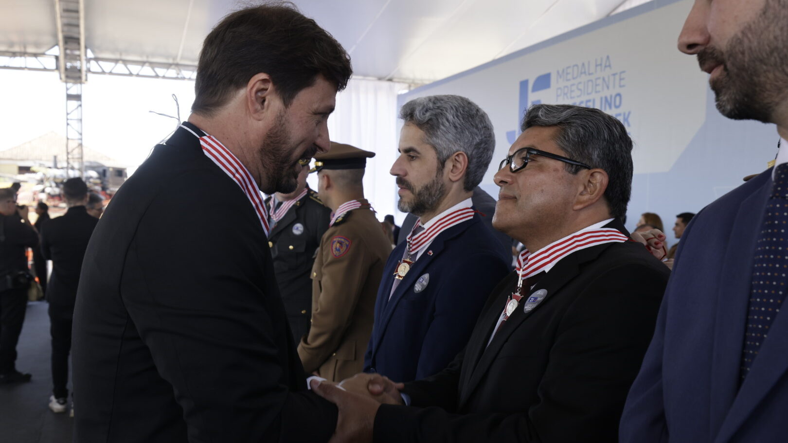 Dois homens de terno e com medalhas no pescoço se cumprimentam com um aperto de mãos durante uma cerimônia. Outros homens de uniforme militar e traje social estão ao fundo.