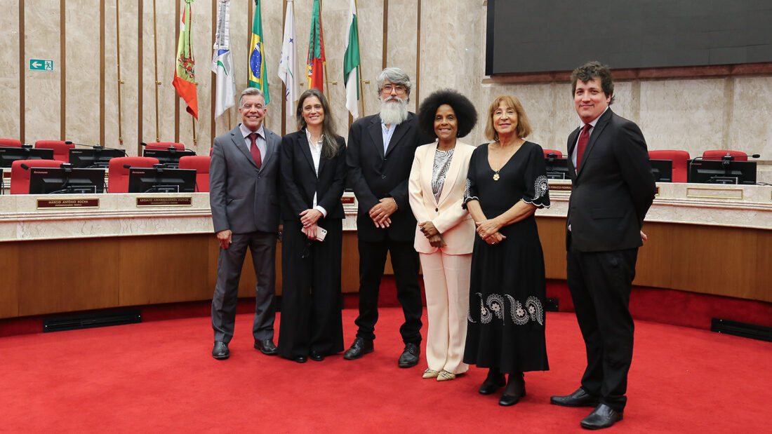 Foto institucional de um grupo de seis pessoas (quatro homens e duas mulheres) em pé, posando juntos em um plenário ou câmara.

O grupo está alinhado sobre um tapete vermelho e, atrás deles, há fileiras de cadeiras e mesas de plenário em tons de vermelho e madeira. No fundo, à esquerda, uma série de bandeiras estão hasteadas. Os trajes são predominantemente formais.