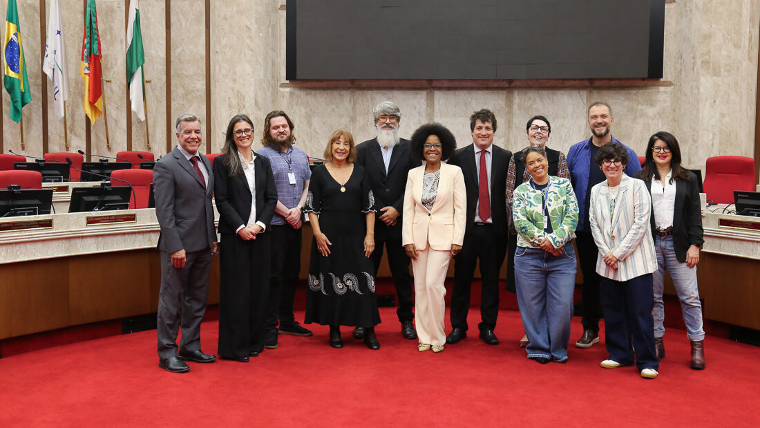 Foto de grupo em um plenário ou câmara, mostrando doze pessoas (homens e mulheres) alinhadas e posando para a câmera.

O grupo é diverso em vestimenta, misturando trajes sociais (ternos escuros, alguns com detalhes vermelhos) e roupas casuais (calças jeans, blazer listrado, camisetas). 

Eles estão em pé sobre um tapete vermelho. O fundo é composto por fileiras de cadeiras e mesas de plenário, painéis de madeira e, no canto superior esquerdo, bandeiras hasteadas. Acima, há uma tela de projeção preta.
