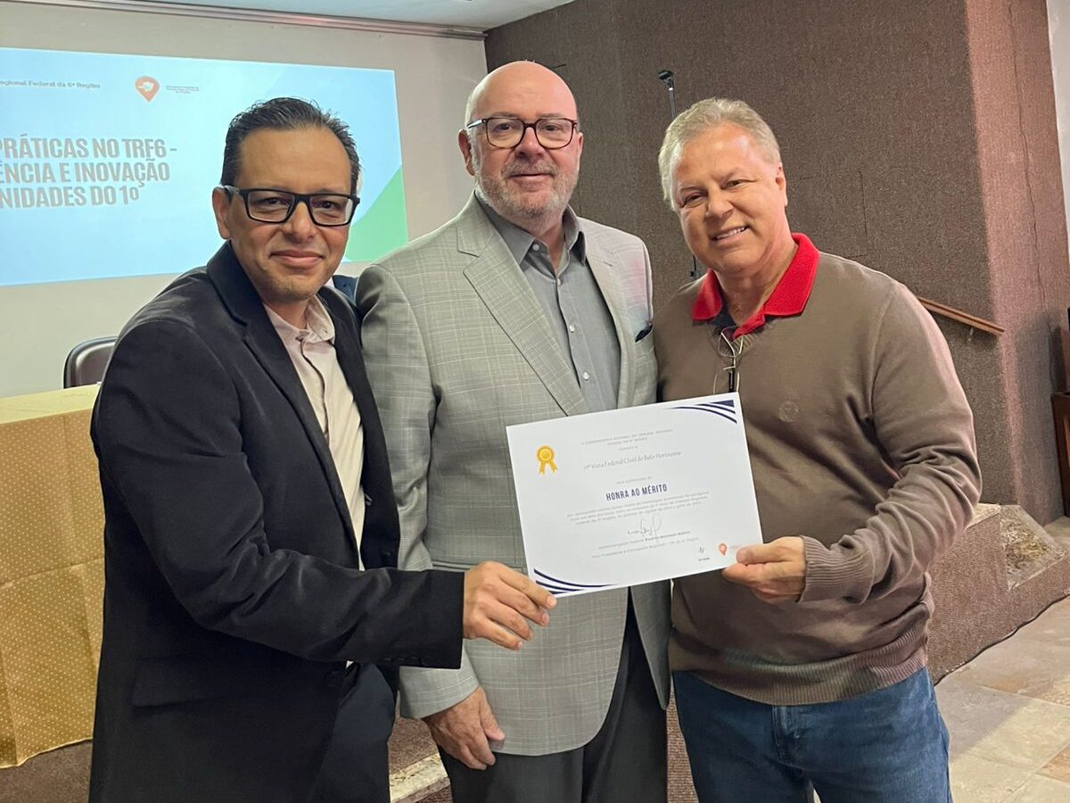 Três homens posam juntos em um palco. O homem à esquerda veste um blazer preto. O homem no centro veste um terno cinza claro. O homem à direita veste uma camisa polo vermelha e um suéter marrom, e ele segura um certificado junto com o homem da esquerda. Uma tela de projeção está visível ao fundo.