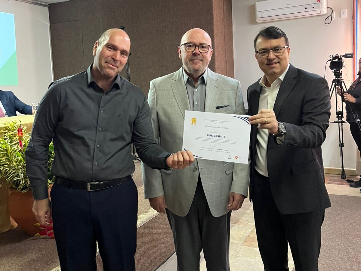 Três homens, vestidos com trajes formais e semi-formais, estão lado a lado em um ambiente de conferência. Dois deles seguram um certificado. Há detalhes de folhagens e equipamentos de gravação no fundo da imagem.