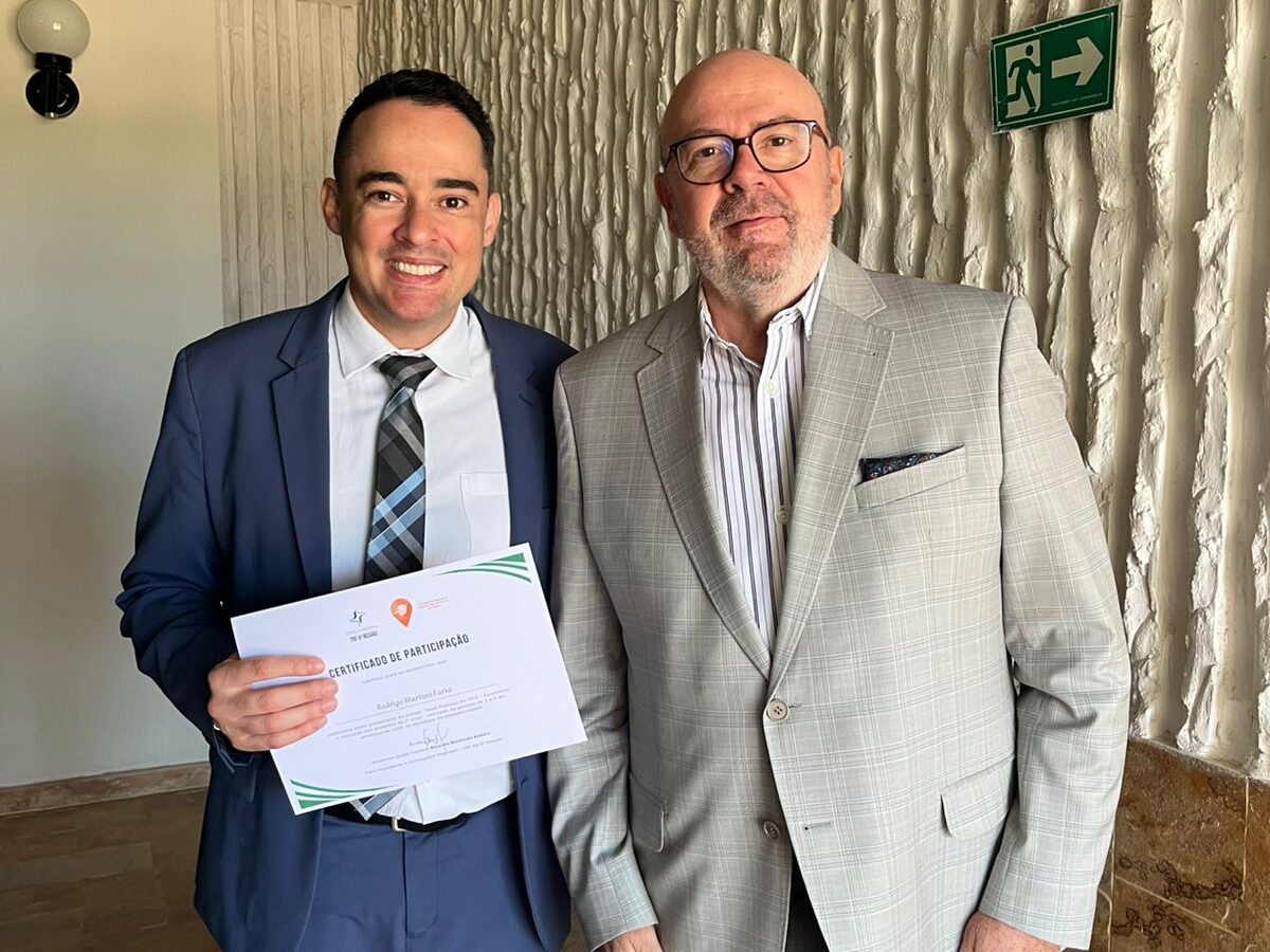 Dois homens posam juntos, vestidos de forma formal e semi-formal, sendo que um deles segura um certificado.