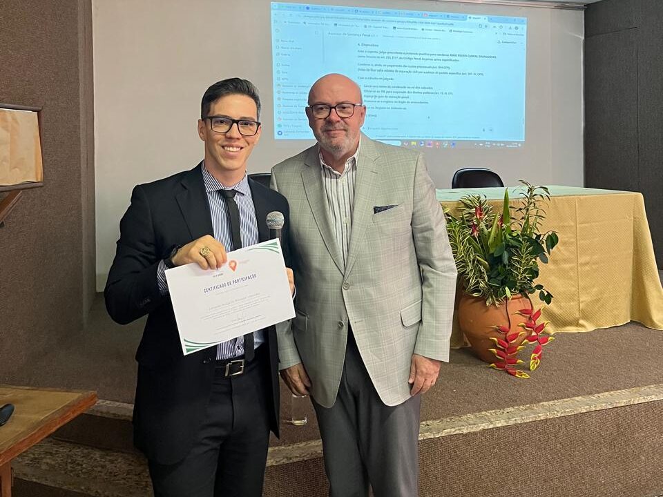 Dois homens posam juntos, vestidos de forma formal e semi-formal, sendo que um deles segura um certificado.
