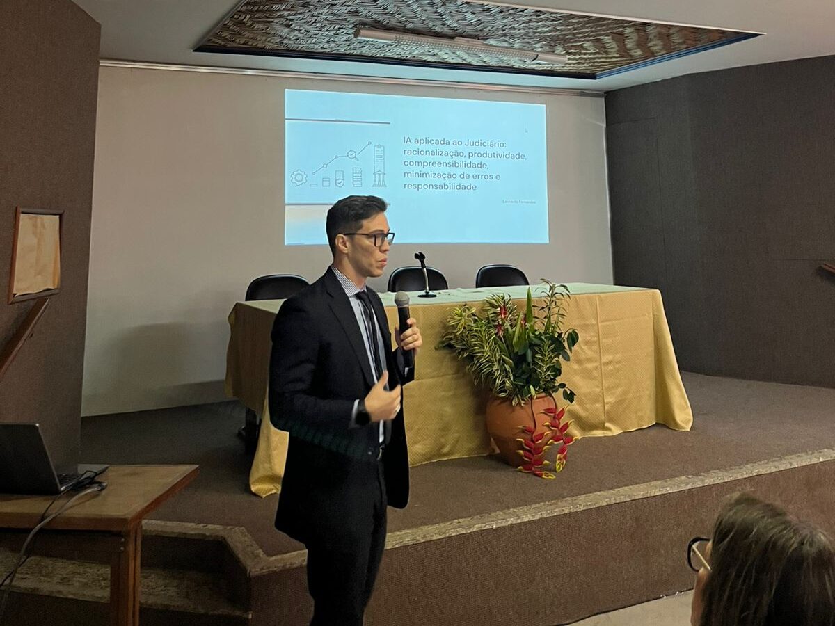 Um homem, vestido de forma formal, está apresentando em um palco ao lado de um púlpito com arranjos de flores.