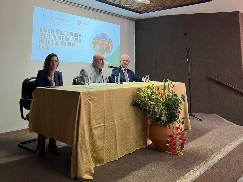 Três pessoas, sendo uma mulher e dois homens, estão sentadas atrás de uma mesa coberta por uma toalha dourada em um auditório. A mulher e um dos homens vestem trajes formais. O segundo homem veste paletó e gravata. À frente da mesa, há um vaso grande de flores. Uma tela de projeção ao fundo exibe o título de uma apresentação.