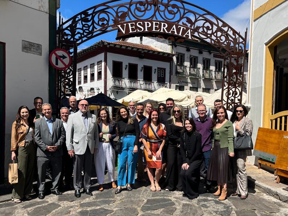 Um grupo grande de pessoas, composto por homens de terno e mulheres vestindo trajes sociais variados, está posando para uma foto em uma rua de paralelepípedos. O grupo está posicionado na entrada de um arco de ferro forjado que tem a palavra "VESPERATA" no topo, e há prédios históricos ao fundo.
