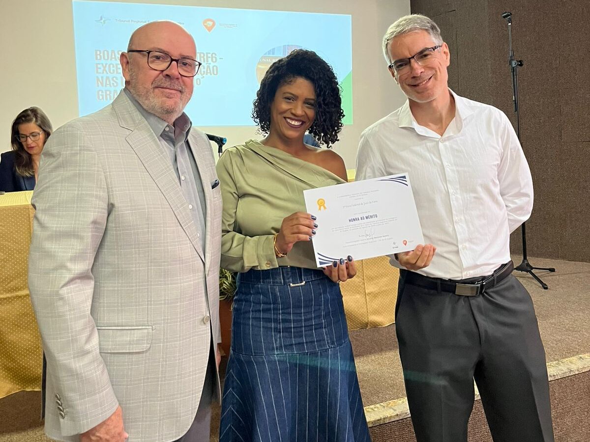 Uma mulher sorridente em destaque, vestindo blusa verde-oliva e saia, recebe um certificado em uma cerimônia. Ela está ladeada por dois homens de terno e camisa social em um palco, com uma tela de projeção visível ao fundo.
