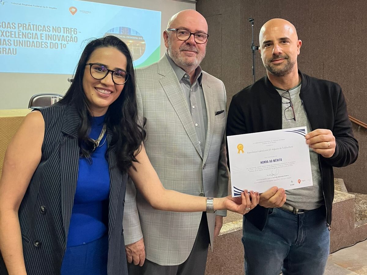 Uma mulher sorridente, com óculos e vestindo uma blusa azul brilhante e colete listrado, posa ao lado de dois homens. Eles estão segurando um certificado de "Honra ao Mérito". O homem no centro veste um terno cinza claro, e o homem à direita veste uma jaqueta preta.