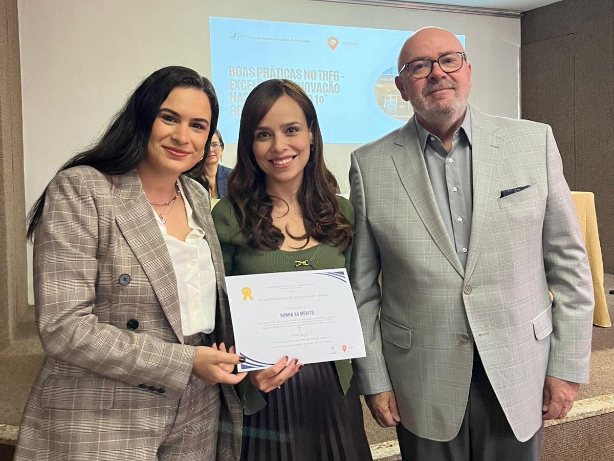 Duas mulheres sorridentes e um homem posam juntos. A mulher no centro segura um certificado e veste uma blusa verde-oliva. A mulher à esquerda veste um blazer xadrez marrom. O homem à direita veste um terno cinza claro. Eles estão em um palco durante um evento.