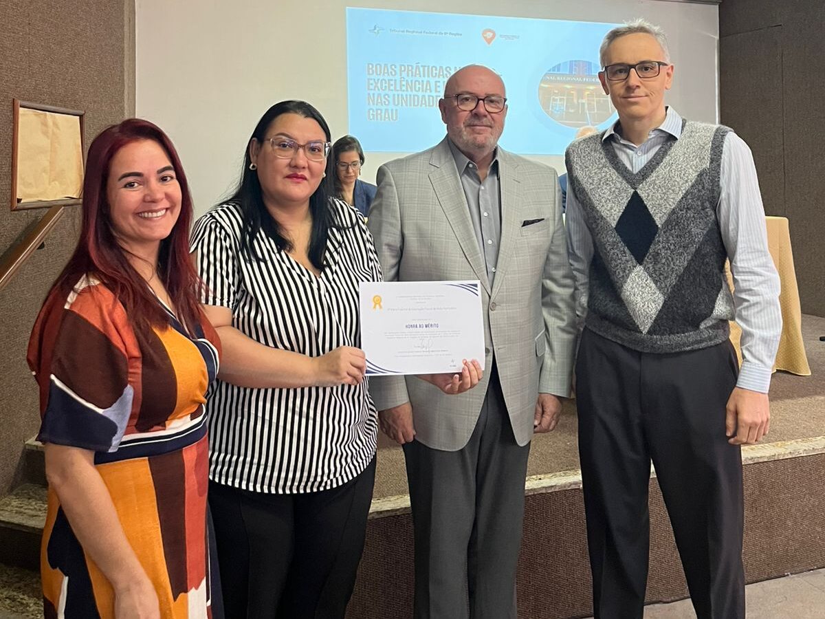 Duas mulheres e dois homens posam juntos em um palco. A mulher à esquerda veste um vestido com padrões coloridos em tons de laranja e marrom. A mulher ao lado dela segura um certificado e veste uma blusa listrada em preto e branco. O homem de terno cinza está ao centro. O homem à direita veste uma camisa social e um colete xadrez em tons de cinza. Há uma tela de projeção visível ao fundo.