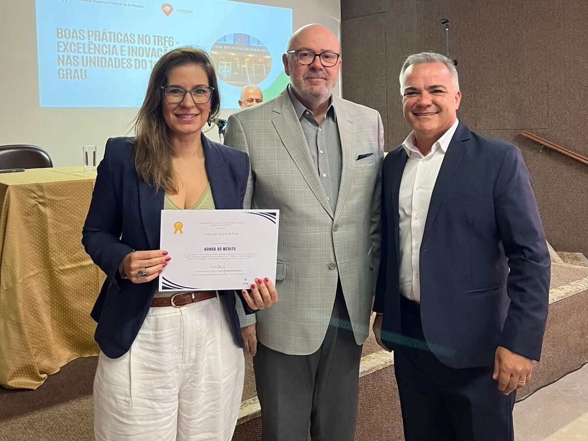 Uma mulher e dois homens estão em um palco de conferências. A mulher, com óculos, está à esquerda segurando um certificado e veste um blazer azul-marinho sobre calças brancas. O homem no centro veste um terno cinza claro. O homem à direita veste um terno azul-escuro com camisa branca. Uma tela de projeção aparece ao fundo.
