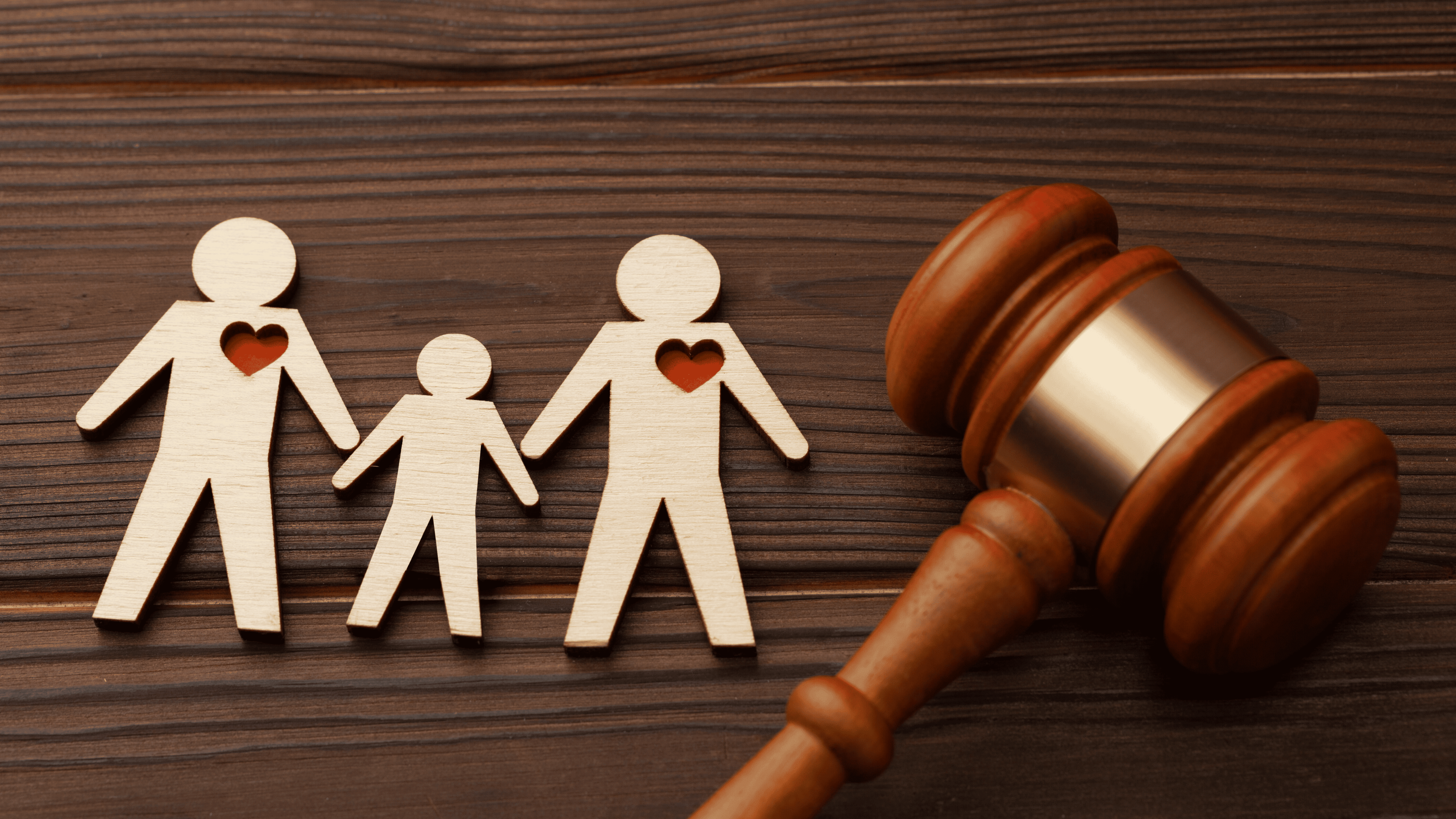 A imagem mostra objetos dispostos sobre uma superfície de madeira escura com veios. No lado esquerdo, estão três silhuetas humanas estilizadas, recortadas em madeira clara, representando uma família: duas figuras adultas maiores e uma figura infantil menor no centro. Cada uma das figuras adultas tem um coração recortado no centro do peito, na cor vermelha. No canto inferior direito, em primeiro plano, encontra-se um martelo de juiz de madeira escura com detalhes em madeira mais clara no topo.