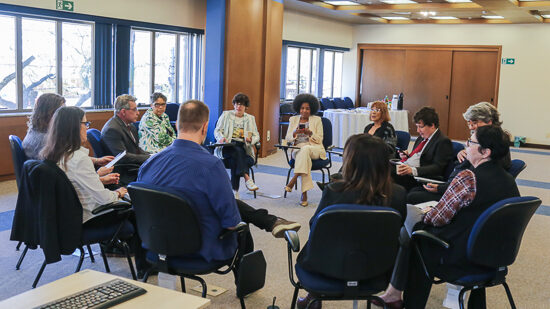 Foto que registra um círculo de debate em um ambiente interno bem iluminado.

Cerca de doze pessoas (homens e mulheres) estão sentadas em cadeiras de escritório pretas com detalhes azuis, dispostas em círculo. A maioria das pessoas está olhando para o centro, com alguns segurando papéis ou documentos.

O fundo é uma sala com grandes janelas que mostram vegetação externa, e paredes com painéis de madeira e detalhes em azul. O chão é coberto por um carpete bege claro. Um teclado de computador é visível no canto inferior esquerdo.