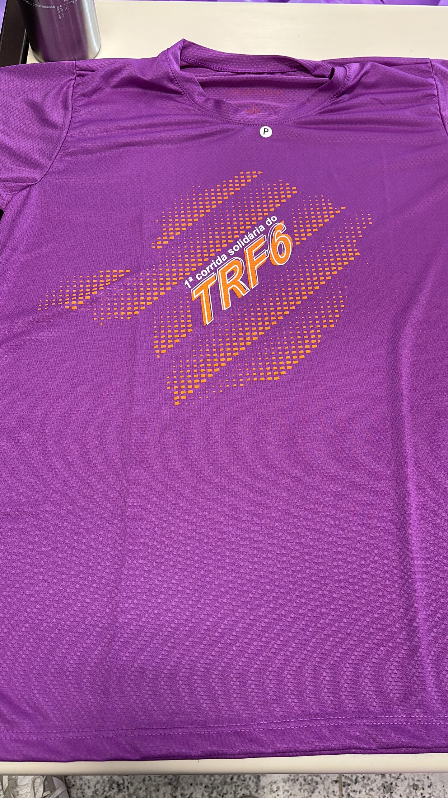 A imagem é uma fotografia em plano fechado de uma camiseta esportiva de cor roxa, possivelmente de tecido tecnológico, que está esticada sobre uma superfície. A camiseta possui uma estampa centralizada na altura do peito, com o texto "1ª corrida solidária do TRF6" em letras laranjas. O texto é emoldurado por um design de linhas diagonais e pontilhados em tons de laranja. A cor roxa da camiseta domina a imagem.