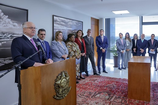Cena de evento formal em ambiente interno, onde um homem de vestes formais (terno e gravata) fala em um microfone atrás de um púlpito de madeira com o brasão da República. Um grupo de outras 11 pessoas, incluindo homens e mulheres em trajes formais e executivos, está posicionado atrás dele. O ambiente possui um tapete estampado, janelas amplas com vista externa e quadros na parede.