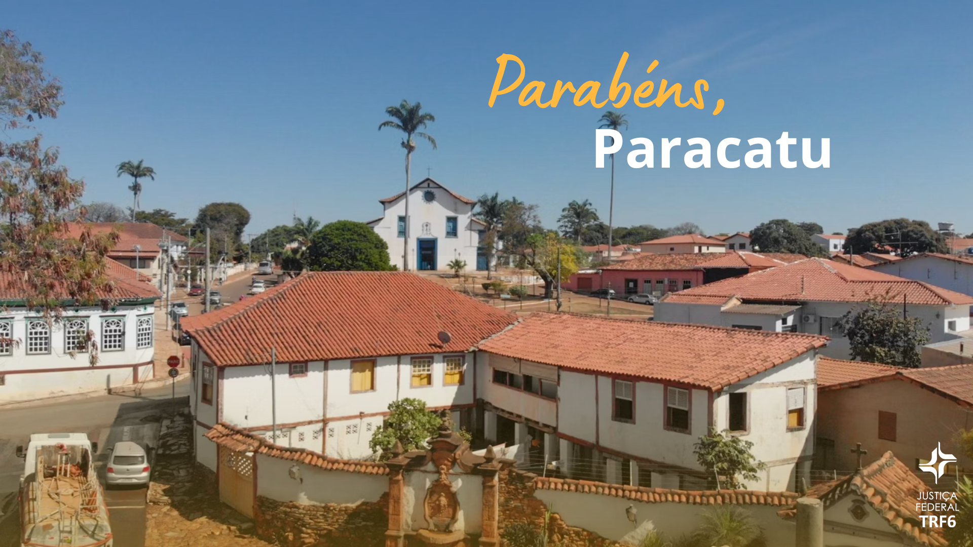 A imagem é uma fotografia aérea de uma paisagem urbana histórica sob um céu azul. O primeiro plano é dominado por telhados de cerâmica vermelha de casas antigas e muros brancos. No centro, no alto, destaca-se uma igreja branca com detalhes azuis. A paisagem urbana mistura edifícios antigos, ruas e vegetação, incluindo palmeiras. No canto inferior direito, há um logotipo sutil da "JUSTIÇA FEDERAL TRF6". Sobre a foto, há uma mensagem de texto em destaque, com as palavras "Parabéns, Paracatu", sendo "Parabéns" em amarelo cursivo e "Paracatu" em branco.
