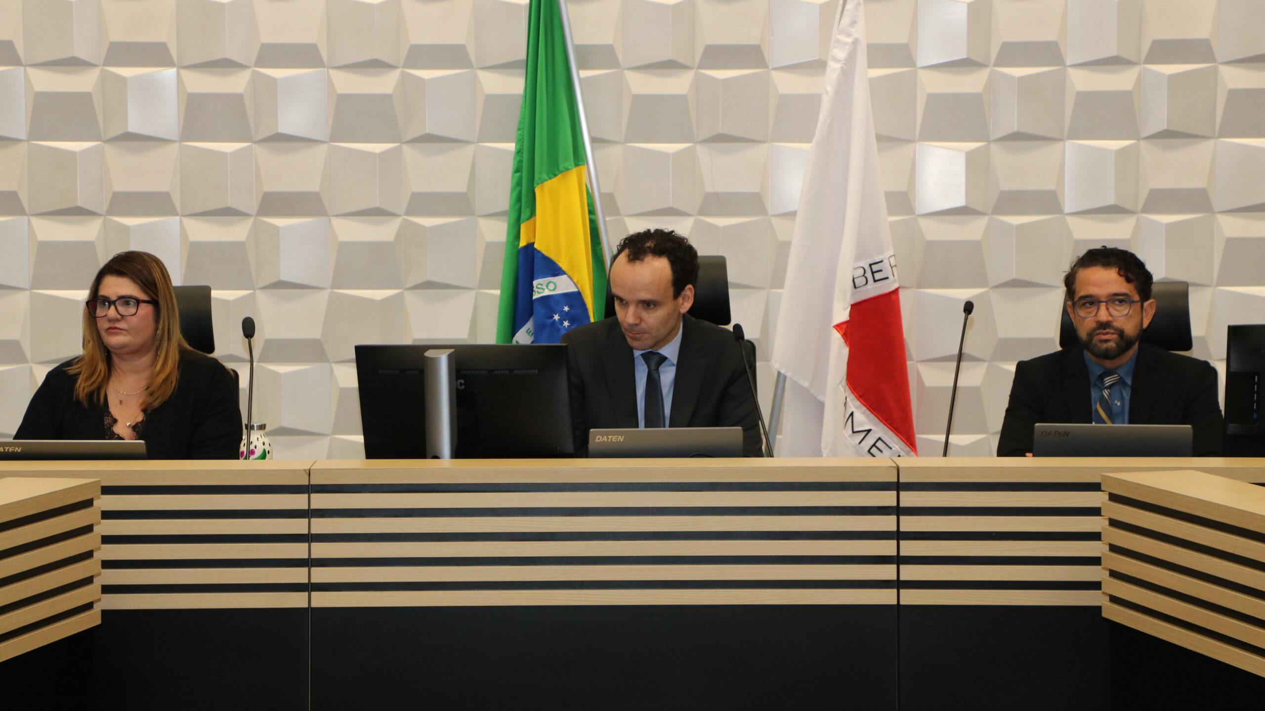 Três pessoas (dois homens e uma mulher) estão sentadas em uma mesa de conferência formal. O homem no meio está olhando para o centro, enquanto a mulher à esquerda e o homem à direita olham para a frente.

Cada um tem um monitor de computador à frente. Atrás deles, na mesa, estão as bandeiras do Brasil e de Minas Gerais. O fundo é uma parede com um padrão geométrico em relevo branco e o revestimento da mesa é de madeira clara com faixas escuras.