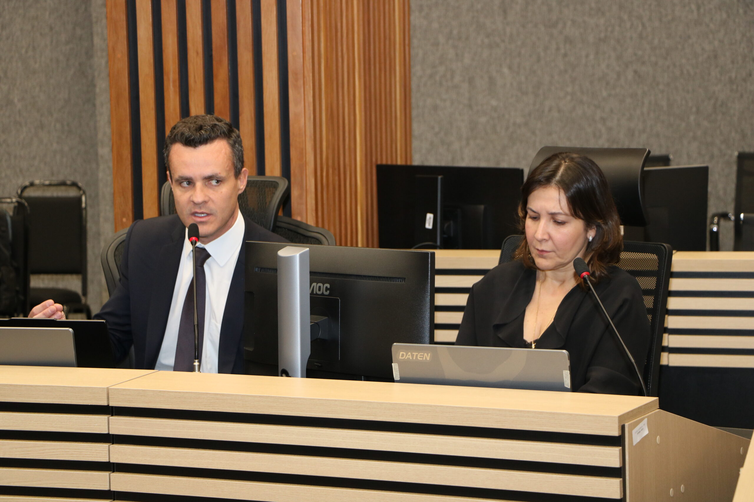 Um homem de terno escuro está falando ao microfone em uma mesa de conferência. Ao seu lado direito, uma mulher de blusa escura está sentada, olhando para baixo, também com um microfone.

Ambos estão em uma sala de reunião formal com mesas de madeira clara e faixas escuras horizontais. Há um monitor de computador entre eles, e as paredes são revestidas com painéis de madeira ripada escura.