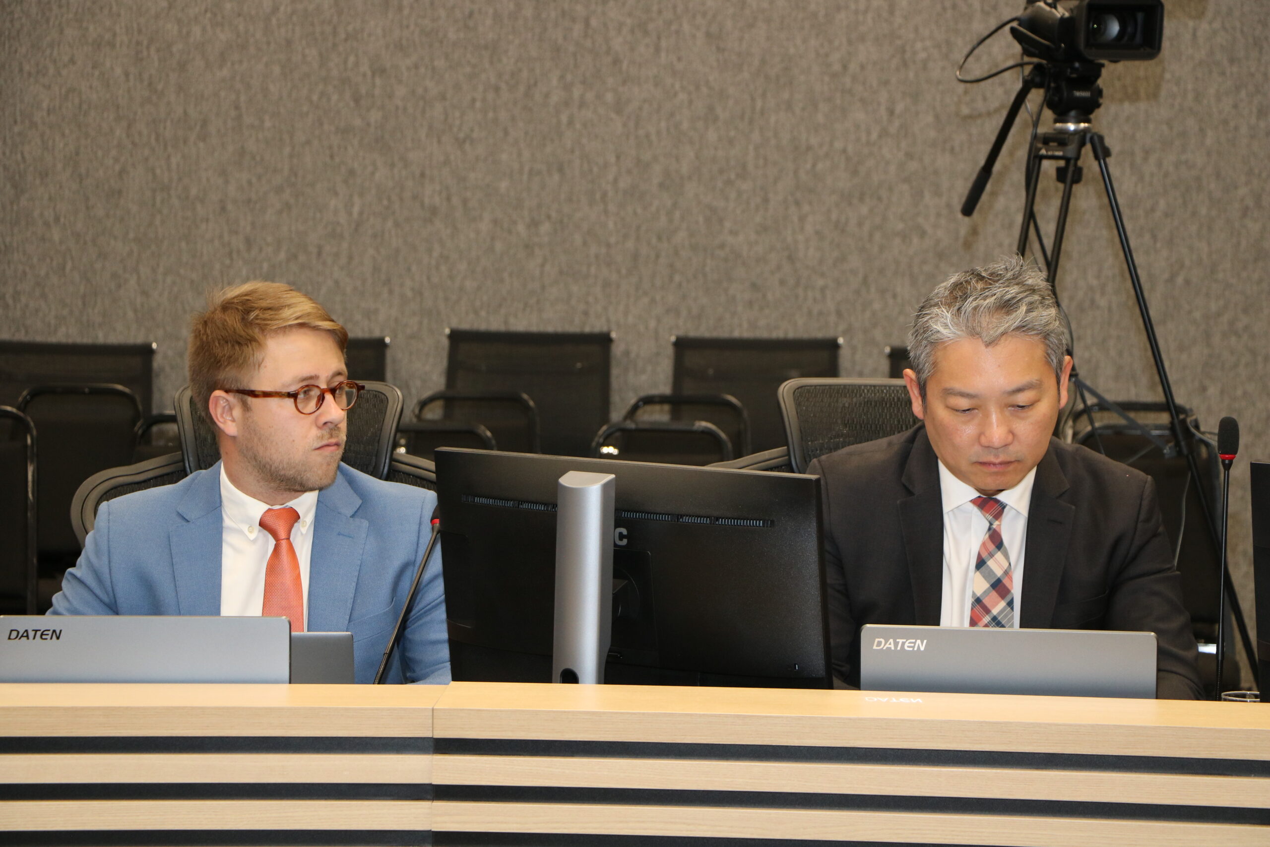 Dois homens estão sentados em uma mesa de conferência formal, cada um com um monitor de computador à sua frente.

O homem à esquerda veste um blazer azul claro com gravata laranja e olha para o lado. O homem à direita veste um terno escuro com gravata xadrez e olha para baixo, em direção ao monitor. O fundo é cinza, com cadeiras pretas.