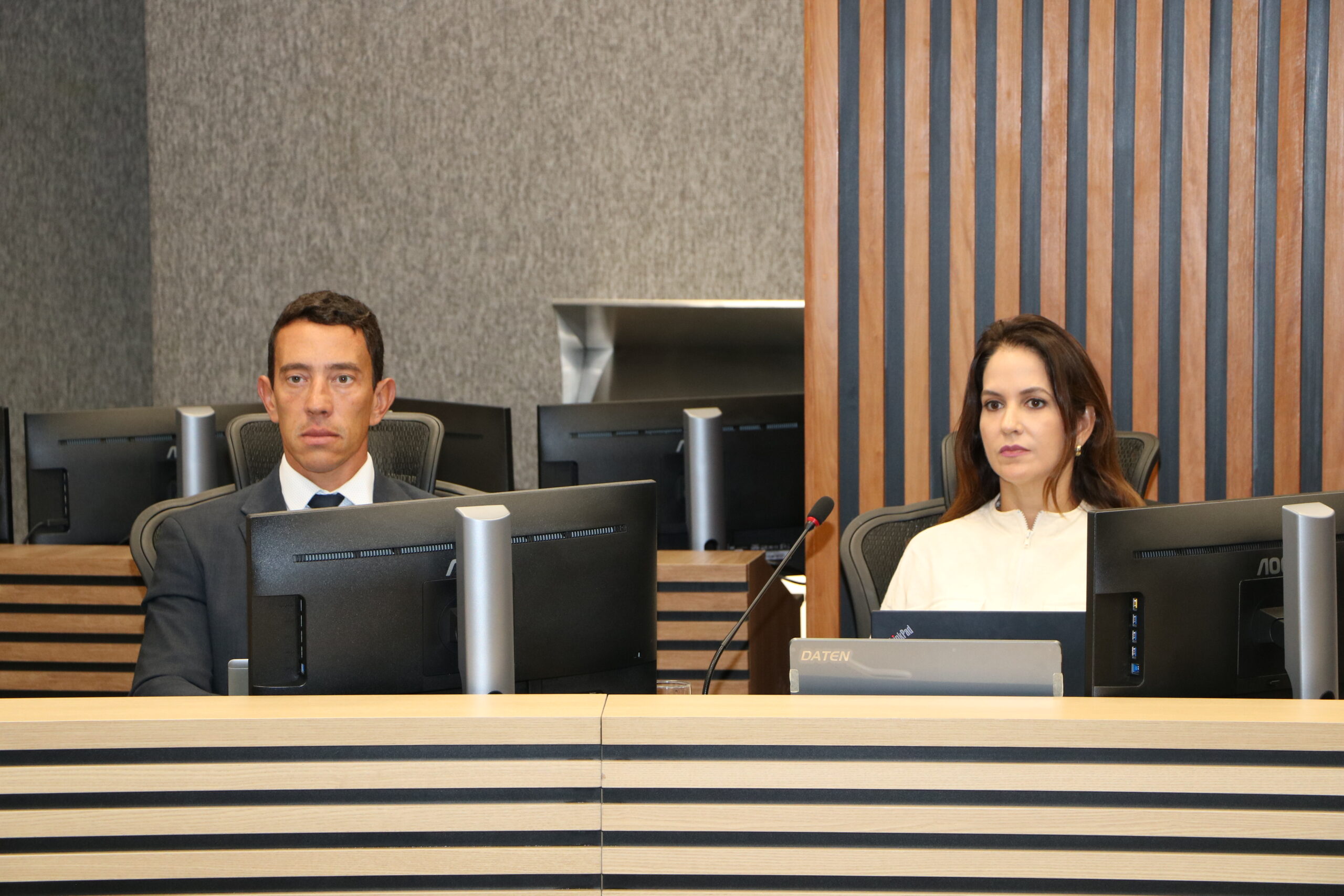 Um homem de terno cinza e uma mulher de blusa branca estão sentados lado a lado em uma mesa de conferência formal. Cada um tem um monitor de computador à sua frente. O ambiente possui um revestimento de madeira ripada escura nas paredes, e outros monitores são visíveis ao fundo.