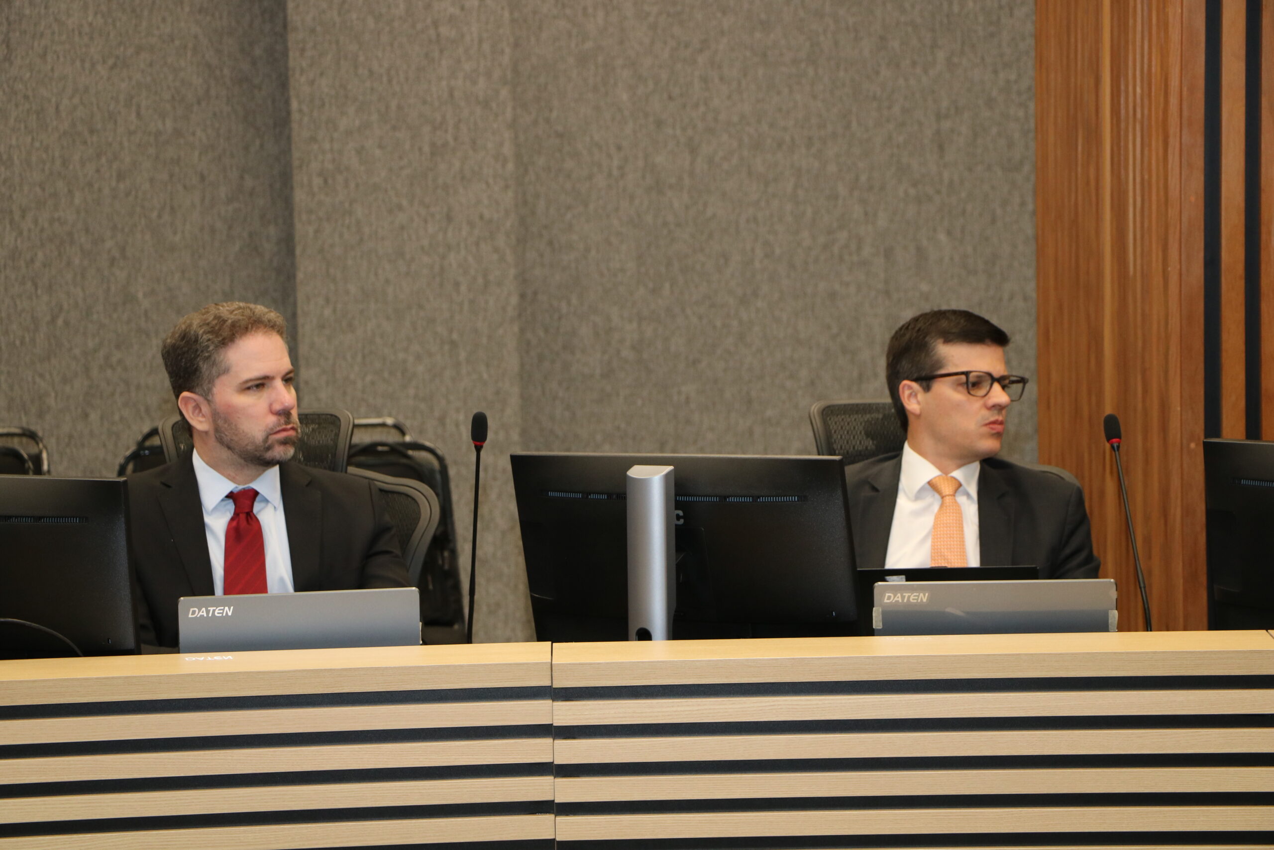 Dois homens estão sentados lado a lado em uma mesa de conferência formal, cada um com um monitor de computador à sua frente.

O homem à esquerda veste um terno escuro com gravata vermelha e olha para o lado esquerdo. O homem à direita veste um terno escuro com gravata laranja clara e óculos, olhando para o lado direito. O fundo é cinza, com painéis de madeira ripada à direita.