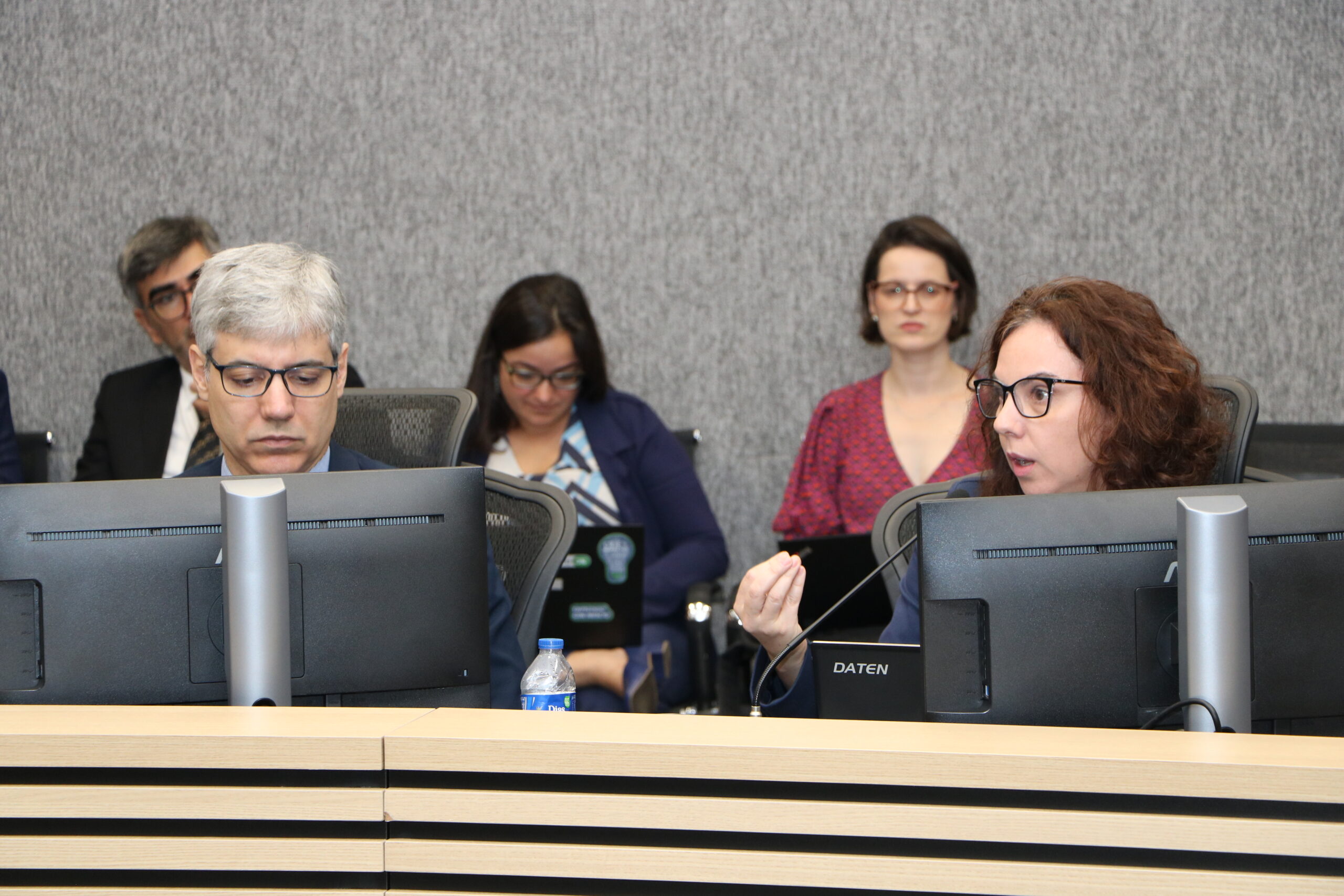 Um homem e uma mulher estão sentados em uma mesa de conferência formal. Em primeiro plano, uma mulher de cabelos cacheados e óculos está olhando para a direita e gesticulando. Ao seu lado, um homem de cabelos grisalhos está olhando para baixo. Todos na frente têm um monitor de computador e uma placa de identificação na mesa. O fundo é de parede cinza, com outros participantes sentados mais atrás.