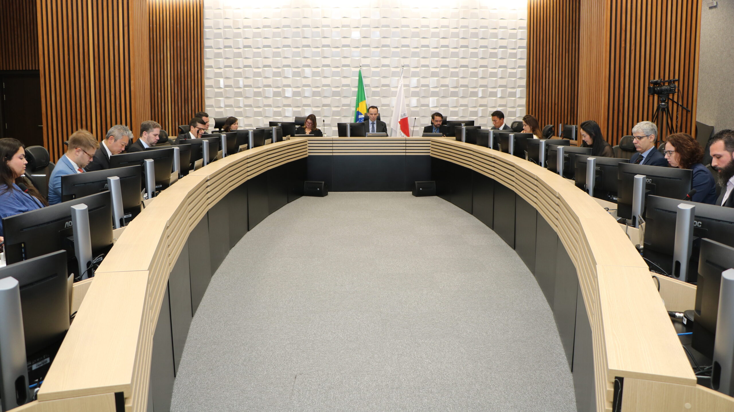 Uma mesa de conferência semicircular com acabamento em madeira clara está disposta em um ambiente formal de sala de reuniões ou plenário.

Várias pessoas estão sentadas ao redor da mesa, voltadas para o centro, e cada uma tem um monitor de computador à sua frente. No centro, contra uma parede com textura tridimensional branca, há uma mesa principal onde um homem está sentado, ladeado pelas bandeiras do Brasil e de Minas Gerais. As paredes laterais são revestidas com painéis de madeira ripada escura.