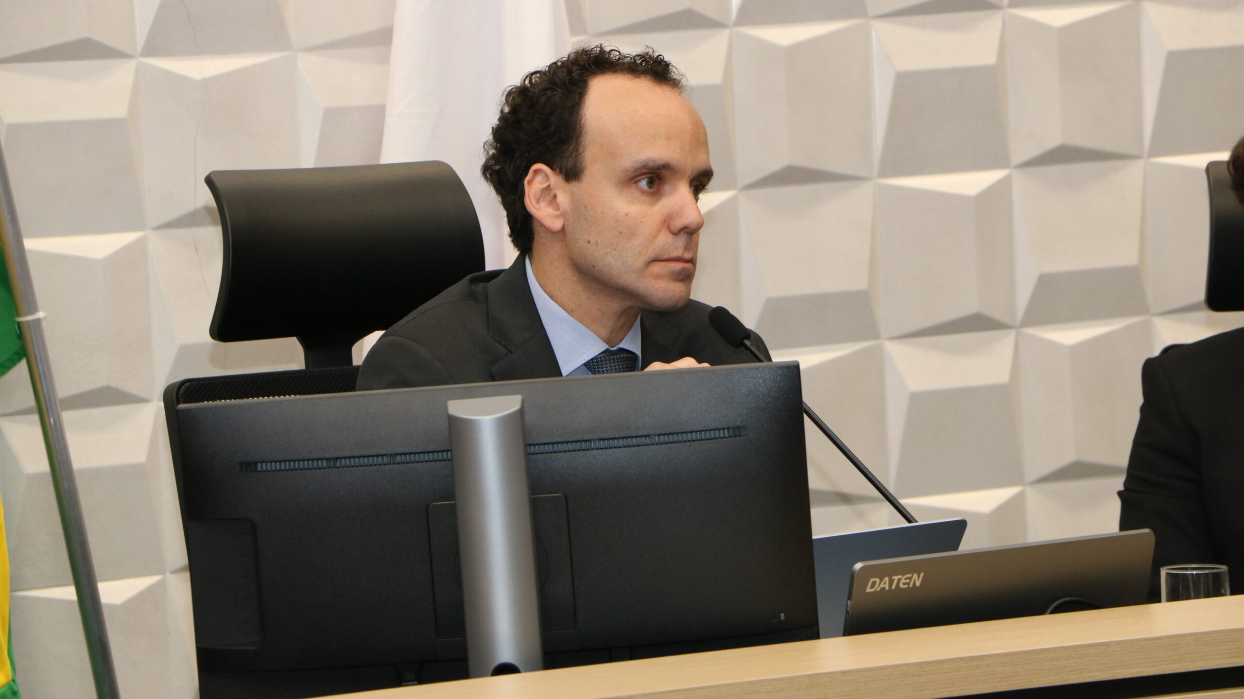 Um homem de cabelos escuros e encaracolados, vestindo um terno, está sentado em uma mesa de conferência ou plenário formal. Um monitor de computador e um microfone estão posicionados na sua frente, sobre uma mesa de madeira clara. O fundo é uma parede com um padrão geométrico em relevo branco e liso, com duas bandeiras visíveis no canto.