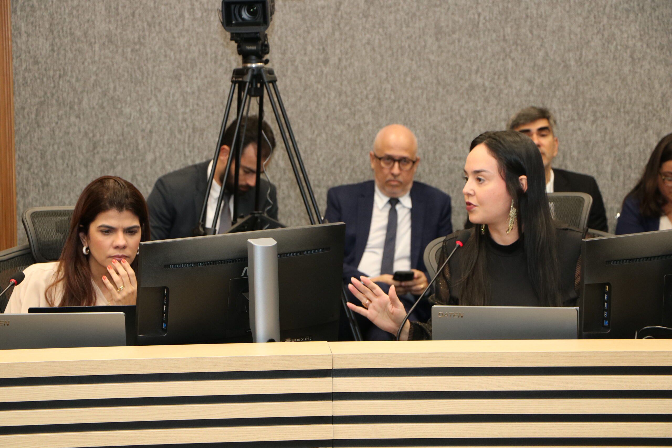 Duas mulheres estão sentadas em primeiro plano em uma mesa de conferência formal, cada uma com um monitor de computador à frente. A mulher à direita está falando e gesticulando com a mão. A mulher à esquerda está olhando para baixo, para o monitor.

No fundo, há três homens sentados. O ambiente possui um tripé de câmera no topo e um revestimento de parede cinza e liso.