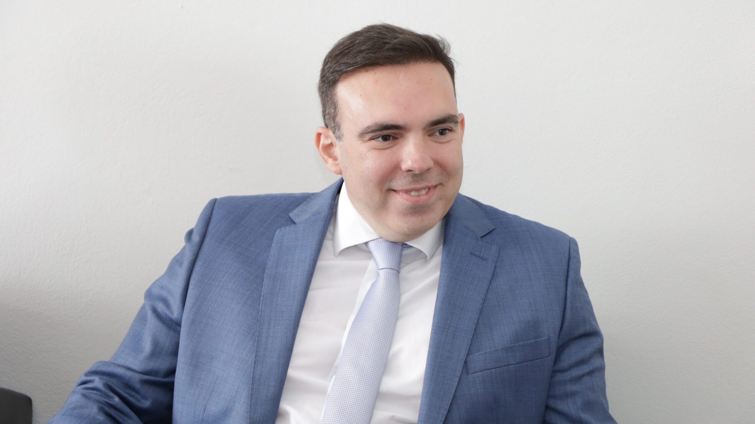 Um homem de cabelo escuro, vestindo um terno azul claro e uma gravata azul-clara estampada, está sentado e sorrindo discretamente, olhando para o lado direito. A foto é um plano médio em um ambiente interno formal, com um fundo de parede branco e liso.