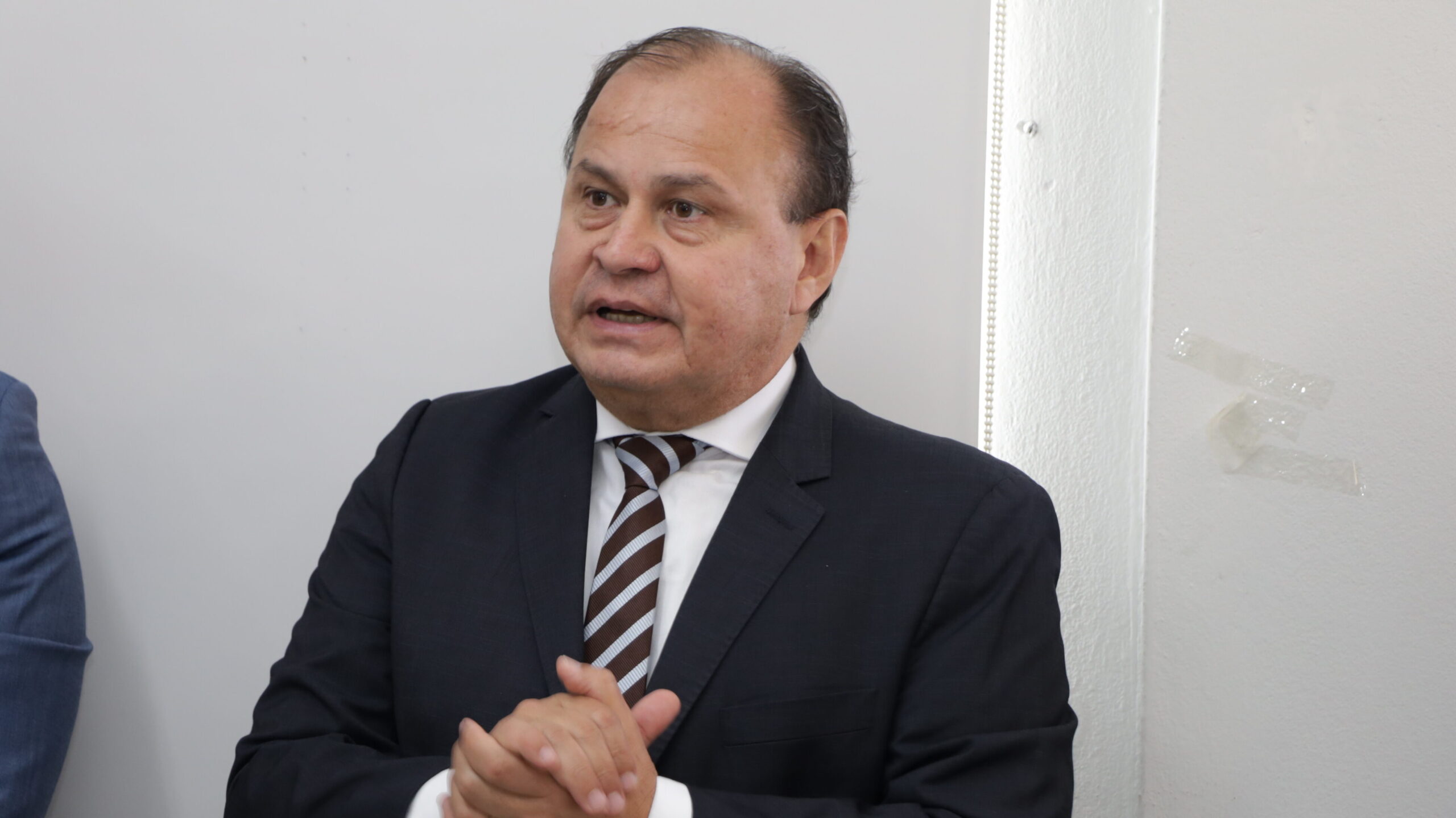 Um homem de meia-idade, vestindo um terno escuro e uma gravata listrada em tons de marrom e branco, está falando ou gesticulando com as mãos unidas. A foto é um plano médio em um ambiente interno formal, com um fundo de parede branca e liso.