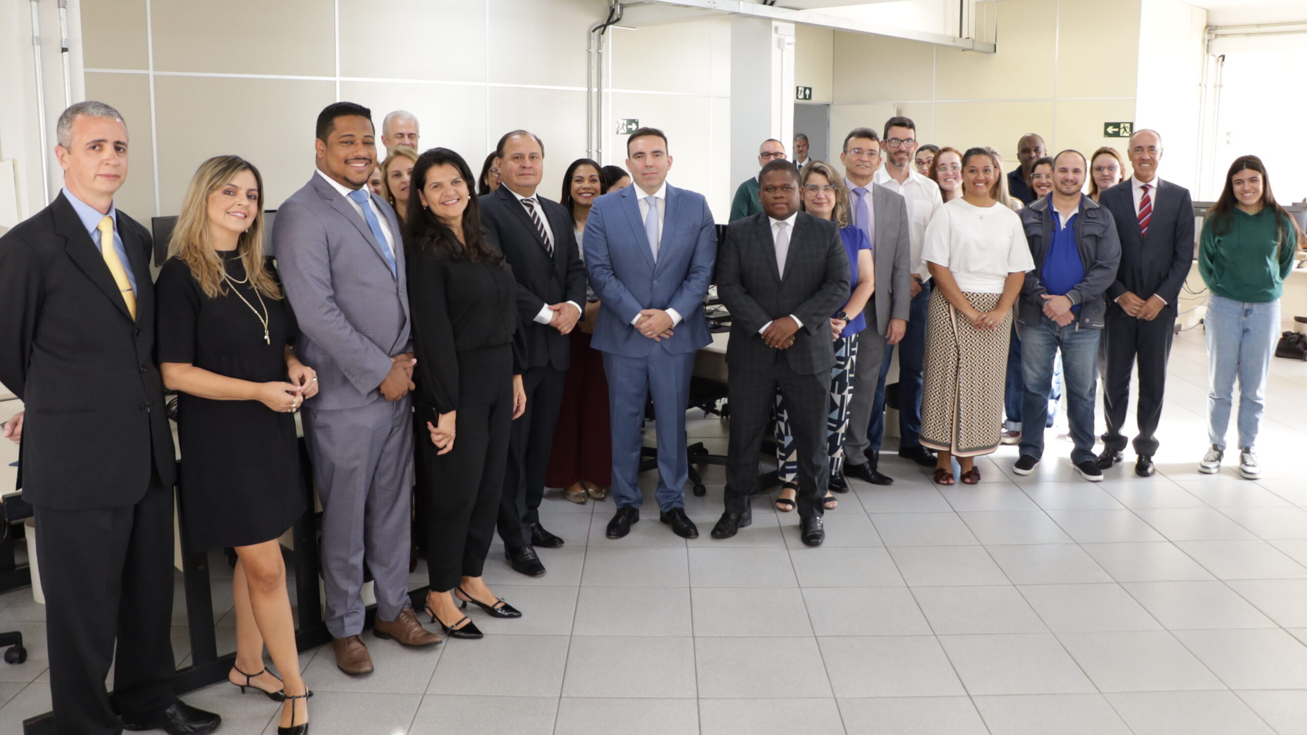 Um grupo de pessoas, incluindo homens e mulheres, está posando para uma foto em um escritório ou sala de reuniões interna. A maioria das pessoas no primeiro plano e no centro veste trajes sociais ou formais, como ternos e vestidos pretos. O grupo está organizado em várias linhas, ocupando o centro da sala com um piso claro e paredes brancas. Computadores e mobiliário de escritório são visíveis ao fundo, assim como sinalizações de saída de emergência.