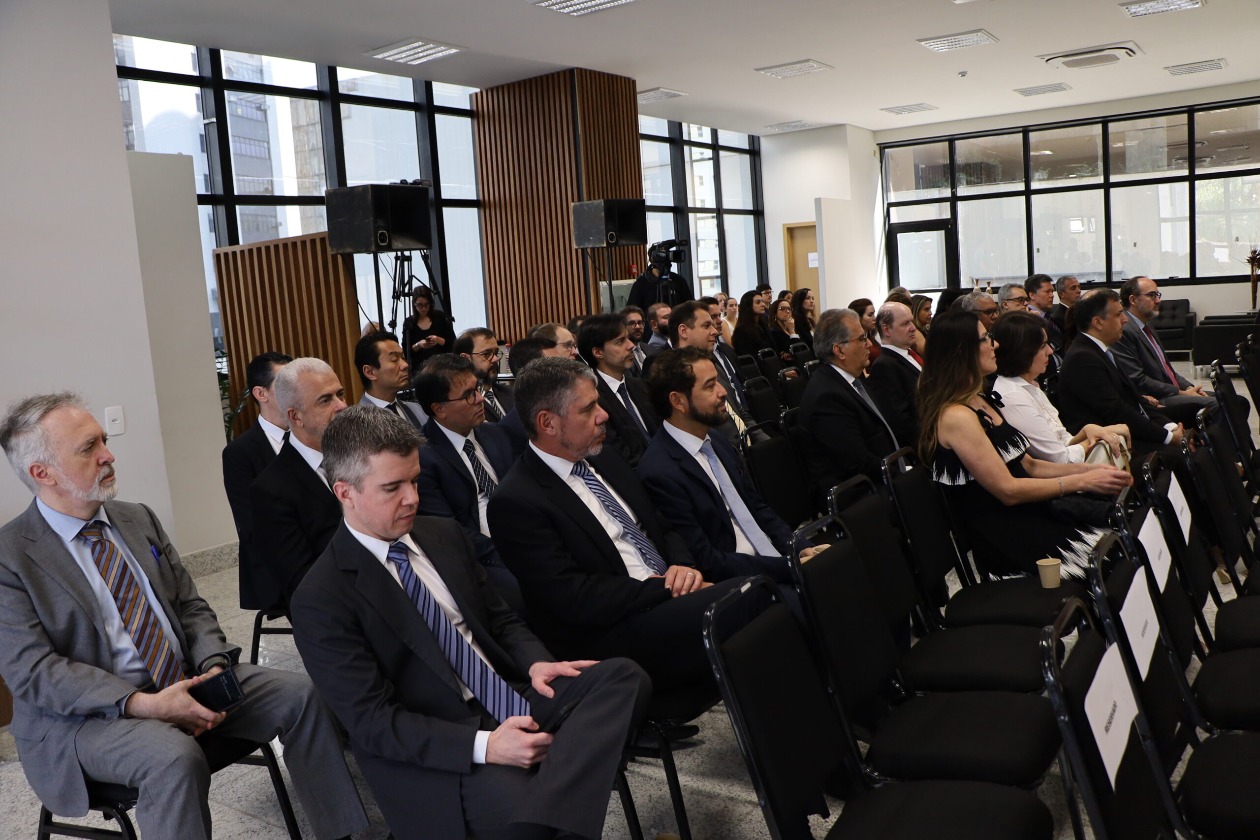 Uma plateia de pessoas em trajes formais está sentada em cadeiras pretas em um auditório ou sala de eventos. A maioria dos participantes é composta por homens de terno e gravata. Eles estão voltados para a frente, assistindo a um evento que não aparece na foto. O local tem paredes claras, grandes janelas com vista para outros prédios e equipamentos de som no fundo.