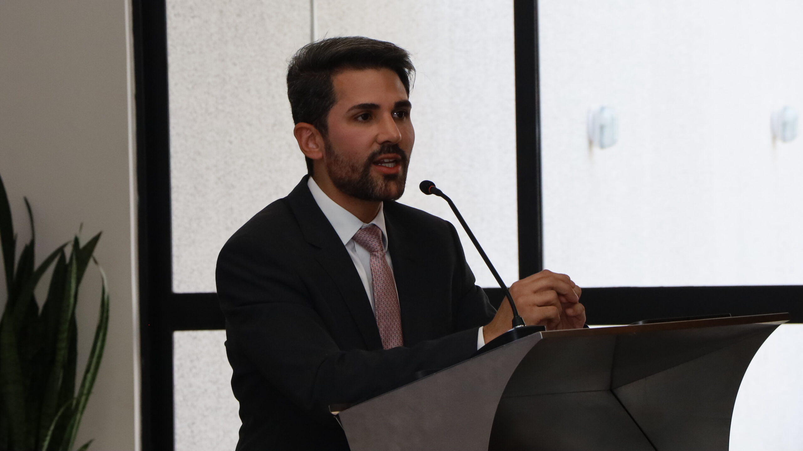 Um homem de barba, vestindo um terno escuro e uma gravata clara com estampa, está falando em um pódio ou púlpito. Ele tem um microfone de haste curvada à sua frente.

A foto é um plano médio que foca no palestrante em um ambiente interno de evento formal, com uma janela grande ao fundo à direita e uma planta verde no lado esquerdo.