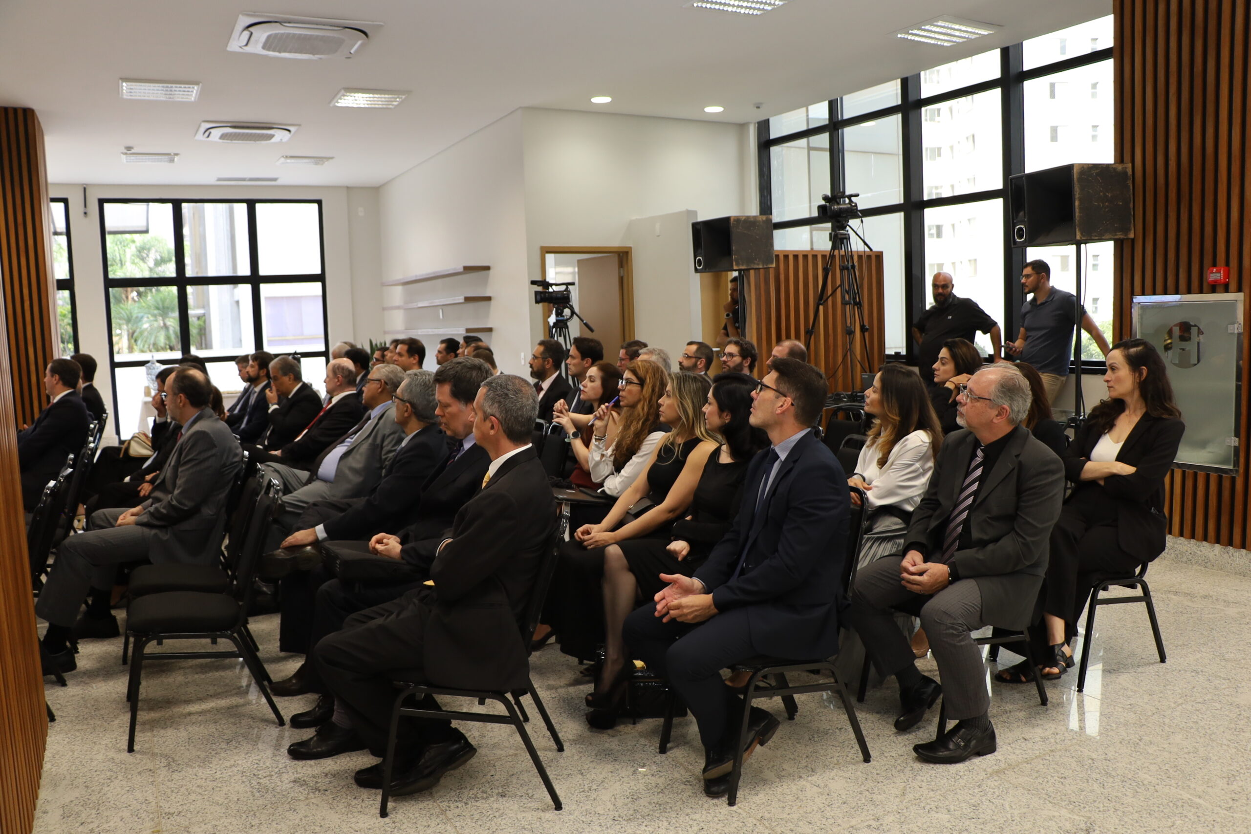 Uma plateia de pessoas em trajes sociais, a maioria de terno, está sentada em cadeiras pretas em uma sala de eventos. Todos estão olhando para a frente e para a esquerda, onde se presume que esteja o palco ou palestrante. O ambiente é formal, com paredes brancas, grandes janelas no fundo e na lateral, e painéis de madeira ripada. Há equipamentos de áudio (caixas de som) e um grupo de pessoas em pé no fundo.