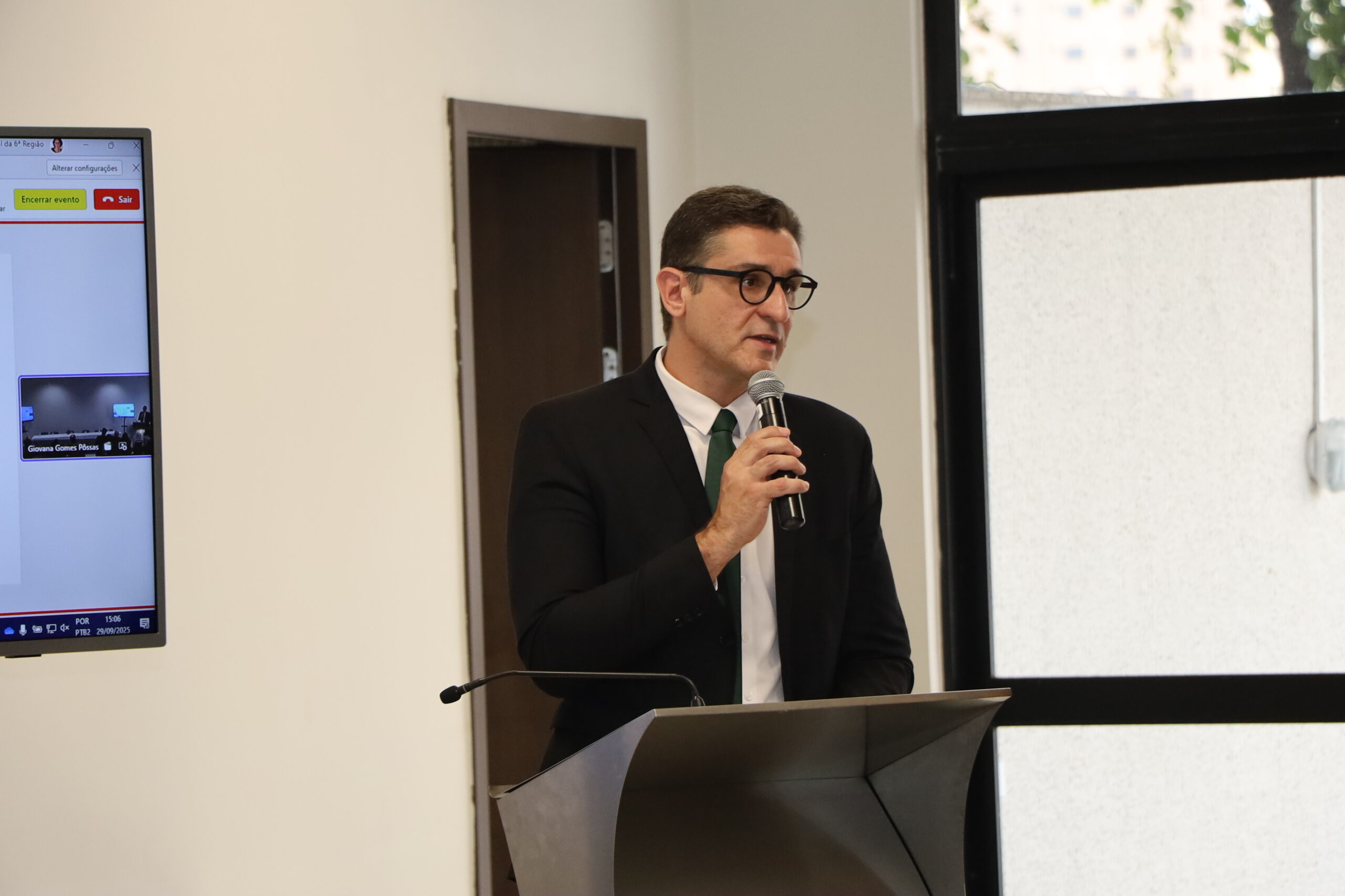 Um homem de óculos, vestindo um terno escuro e gravata, está falando em um pódio ou púlpito cinza, segurando um microfone de mão.

A foto foi tirada em um ambiente interno formal, com uma grande janela à direita e uma porta escura ao fundo. À esquerda, um monitor exibe uma tela com botões e uma visualização de vídeo.