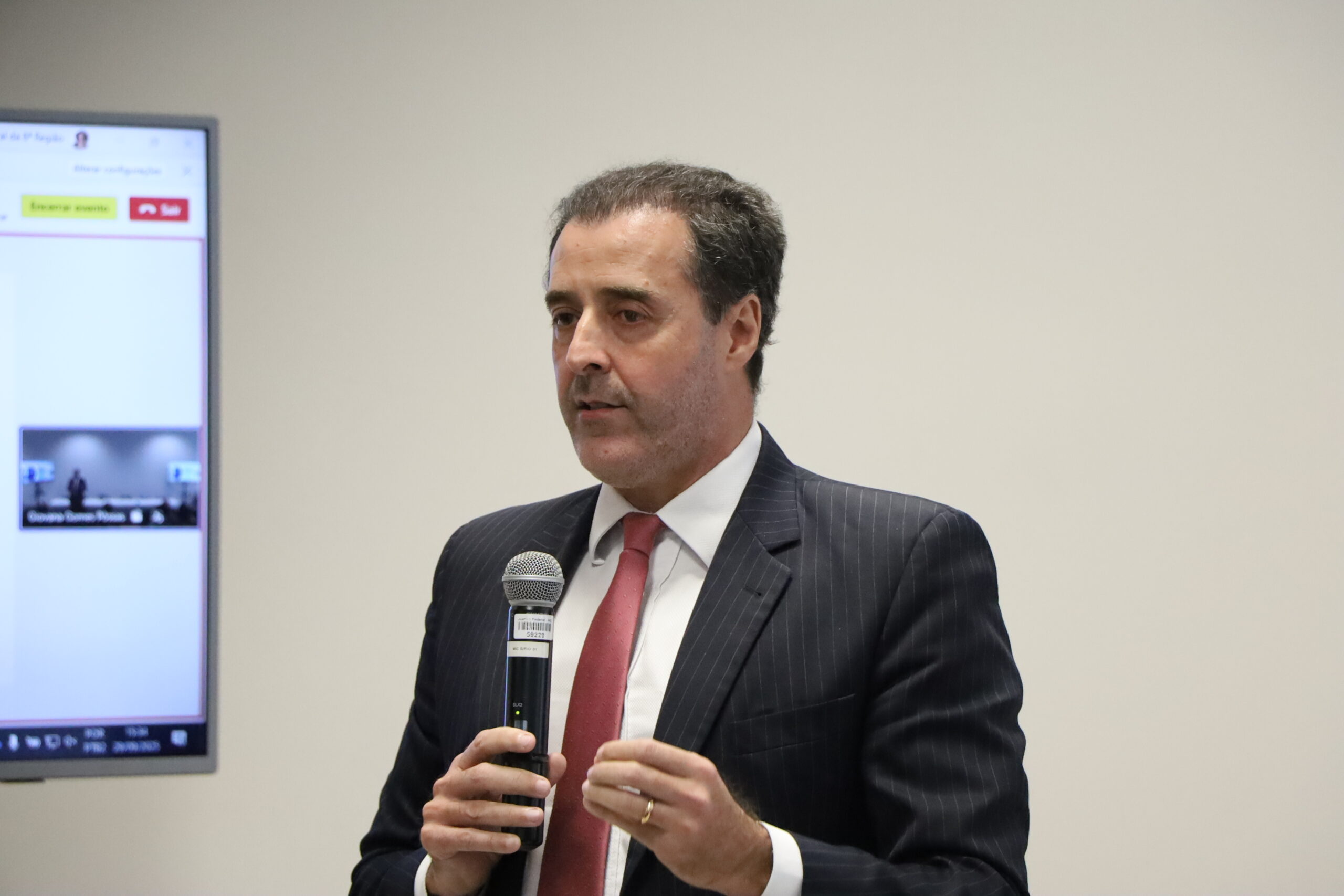 Um homem de meia-idade, vestindo um terno risca de giz escuro e uma gravata vermelha, está falando ao microfone. Ele segura o microfone de mão com as duas mãos e gesticula levemente com os dedos.

A foto é um plano médio do orador em um ambiente interno formal, com um monitor exibindo uma tela de videoconferência no lado esquerdo e um fundo de parede branco e liso.