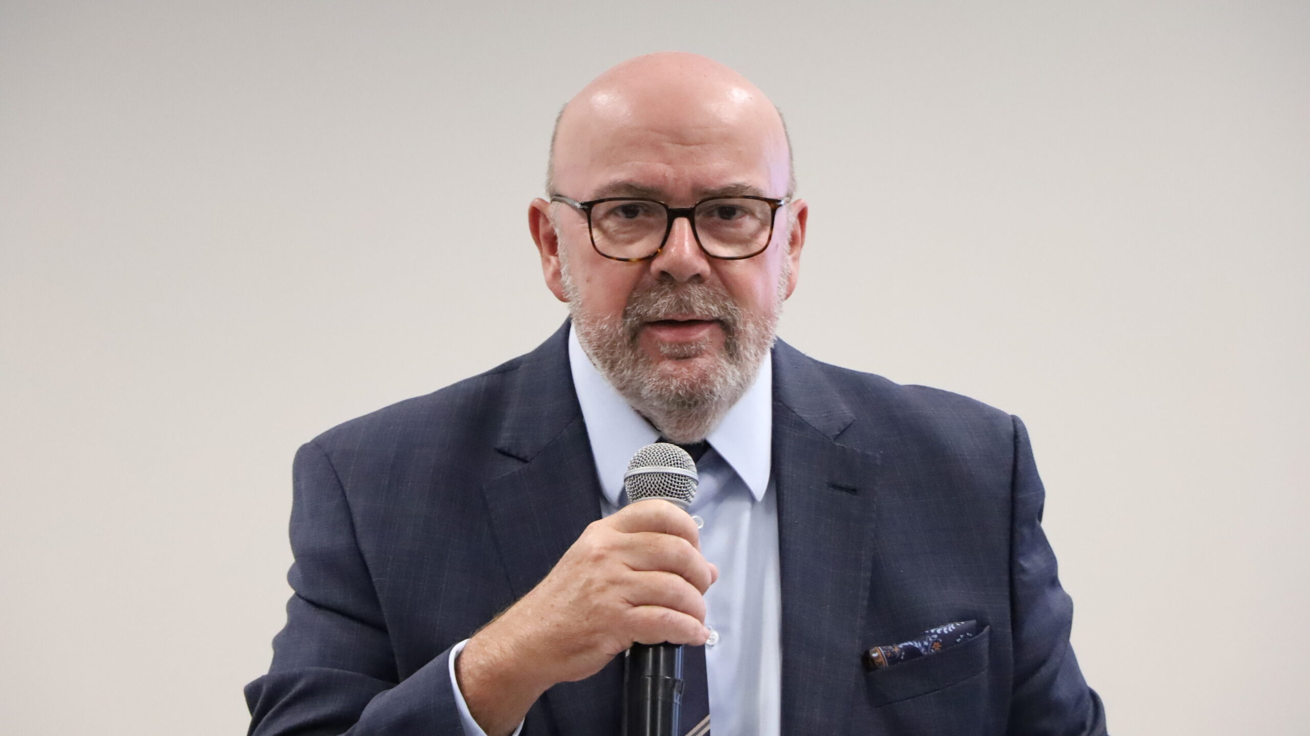 Um homem calvo de barba, vestindo um terno azul marinho e óculos, está falando, segurando um microfone de mão próximo à boca. Ele usa um lenço de bolso estampado. A foto é um plano próximo, focado no rosto e tronco superior do orador, com um fundo liso e claro.