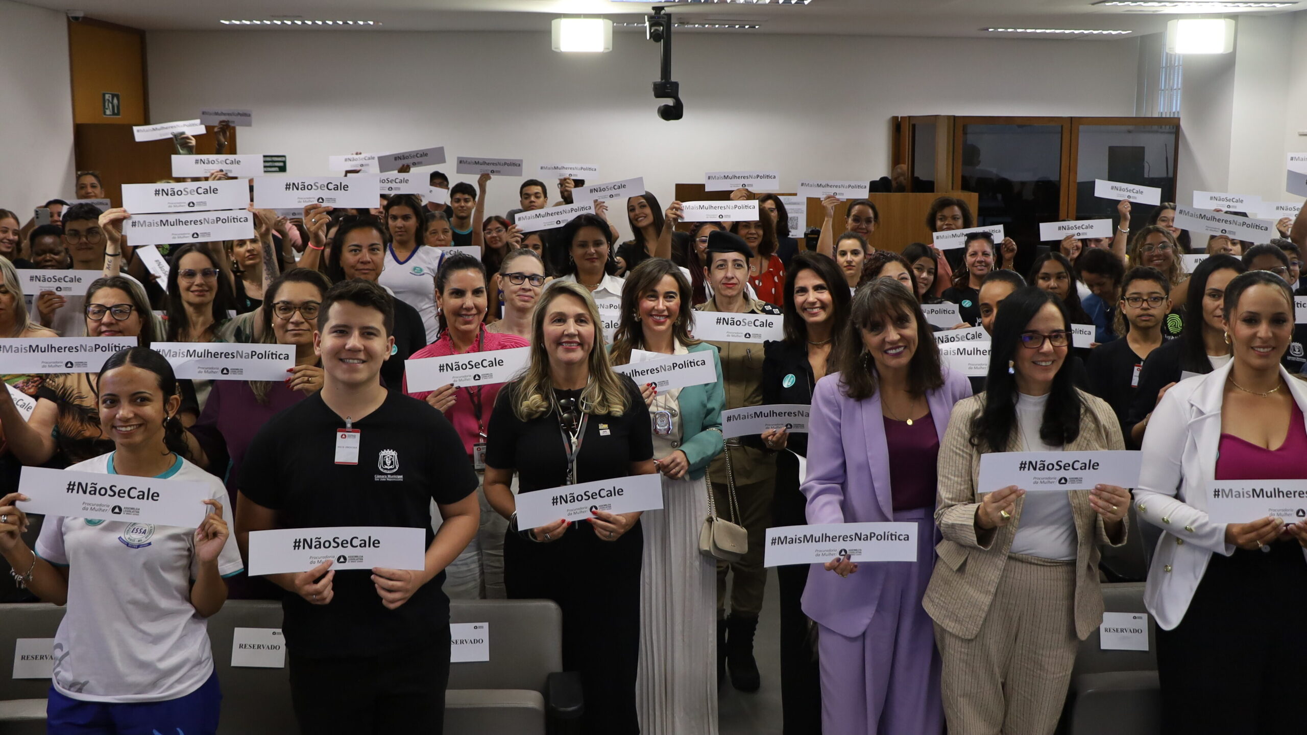 Foto de grupo em um auditório.

Um grande grupo de pessoas (homens e mulheres) está em pé, em fileiras escalonadas. Quase todos estão segurando pequenos cartazes brancos com hashtags como: #NãoSeCale e #MaisMulheresNaPolítica.

O auditório possui assentos escuros e painéis de madeira clara ao fundo.
