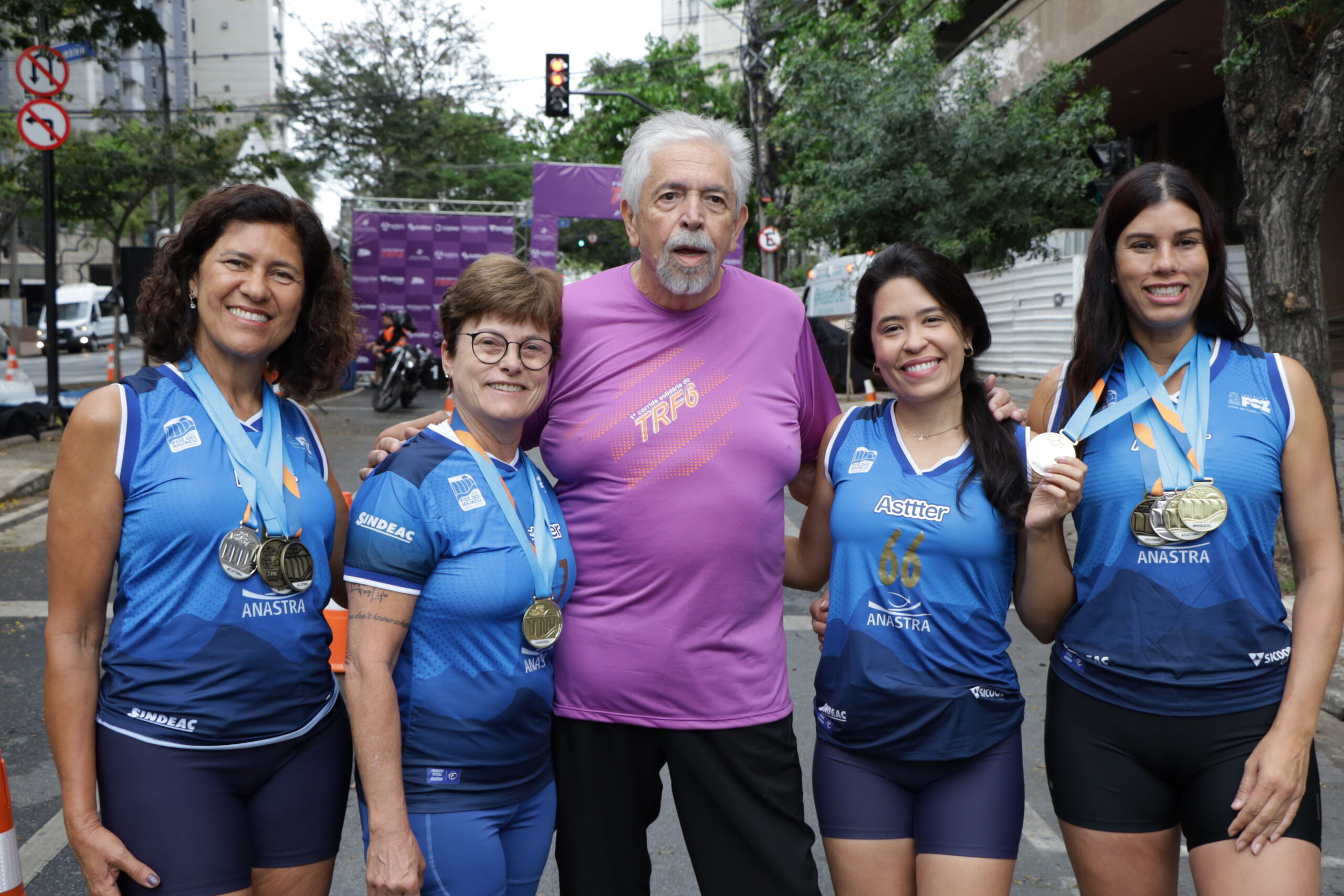 Quatro mulheres em camisetas azuis de corrida, exibindo medalhas, posam ao lado de um homem de camiseta roxa, em uma rua urbana.